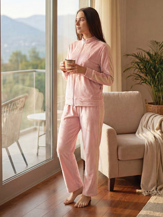 A woman stands in pink pajama bottoms and a matching jacket, holding a mug and looking out a window.