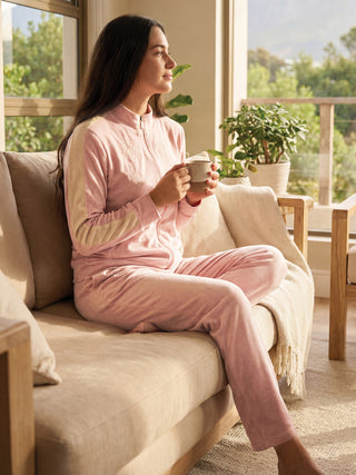 A woman in a light pink velour tracksuit with floral embroidery sits on a couch, holding a mug, looking out a window.