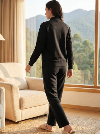 A woman is seen from behind, wearing a black zip-up tracksuit with subtle glitter accents, standing in a cozy room by a window overlooking a scenic view.