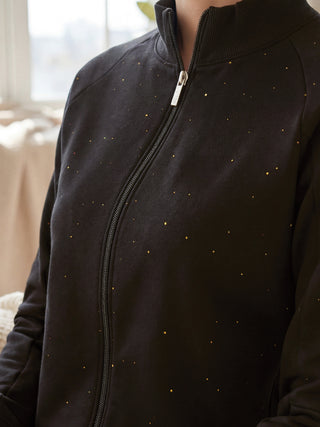 A woman is seen from the chest up, wearing a black zip-up jacket with a subtle gold speckle print.