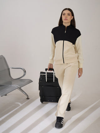 A woman walks with a suitcase wearing a beige tracksuit with black and beige color blocking.