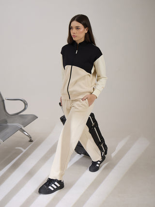 A woman models a beige tracksuit with black and beige color blocking, posed with a suitcase.