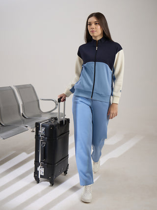 A woman walks with a black suitcase, wearing a colorblock tracksuit in navy, light blue, and cream.