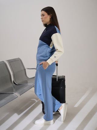 A woman with long brown hair stands next to a black suitcase wearing a blue, cream, and navy color-blocked track suit.