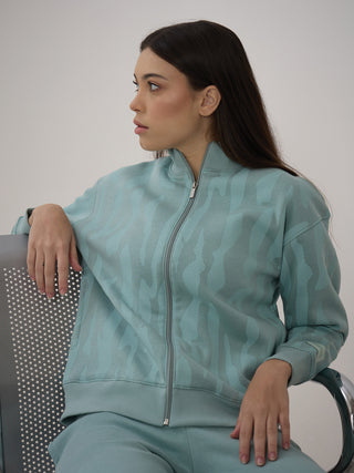 A woman in a mint green printed zip-up jacket and matching pants sits on a chair, looking away from the camera.