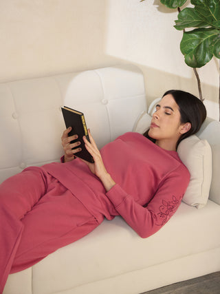A woman is lying on a white couch, wearing a pink sweatshirt with floral embroidery and matching sweatpants, and reading a book.