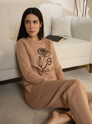 A woman wearing a tan loungewear set with a rose line-art print sits on the floor.