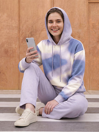 A woman sits on striped steps, wearing a lavender and blue tie-dye hoodie and matching lavender sweatpants, holding a phone.