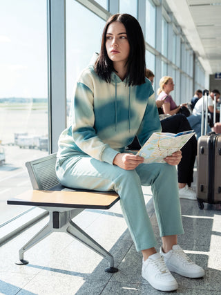 A woman wearing a teal and white tie-dye tracksuit sits in an airport, holding a map.