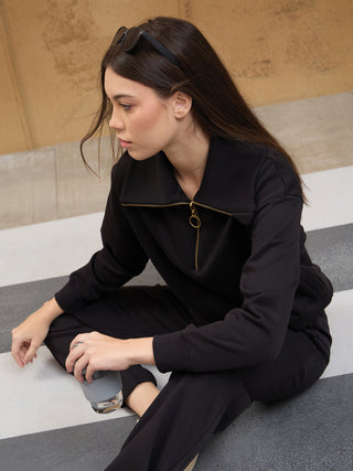 A young woman in black sweatpants and a black zip-up hoodie sits on a striped floor, holding a water bottle.