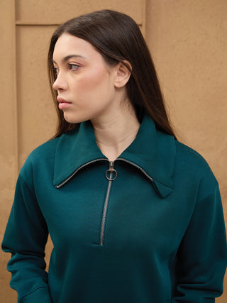 A woman in a teal zip-up sweatshirt and brown pants poses on a brown background.