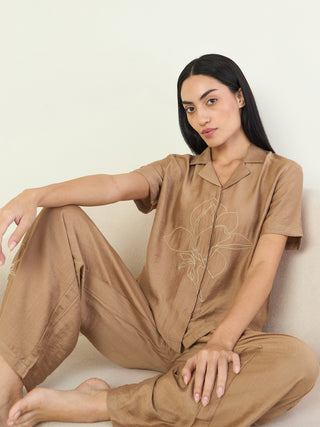A woman sits on a couch wearing a tan silk pajama set with a delicate white floral print on the shirt.