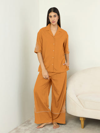 A woman poses in a relaxed stance, wearing a mustard-colored pajama set featuring subtle white embroidery on the sleeves and pant cuffs.
