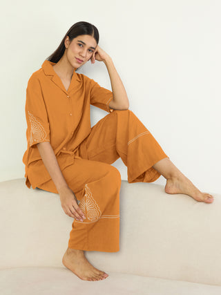 A woman poses in a relaxed stance, wearing a mustard-colored pajama set featuring subtle white embroidery on the sleeves and pant cuffs, unlike the previous image, this one shows her sitting on a couch.
