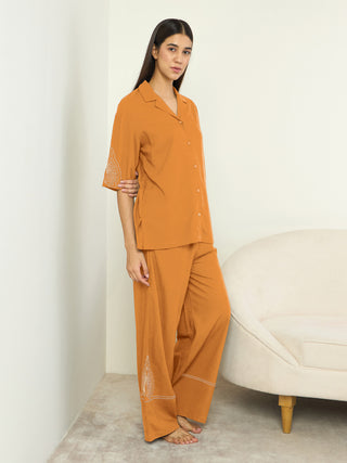 A woman poses wearing a rust-colored pajama set with white embroidery on the sleeves and pant legs, sitting on a cream-colored sofa.