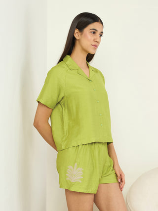 A woman in a lime green pajama set with floral embroidery stands against a white wall.