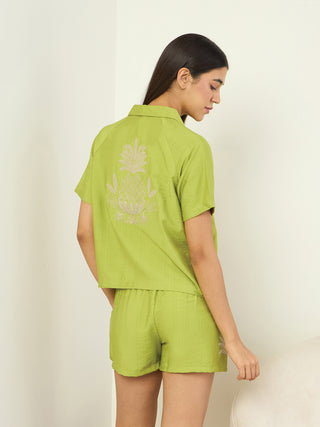 A woman is posing for a product shot of a green pajama set with an embroidered pineapple on the back of the shirt.