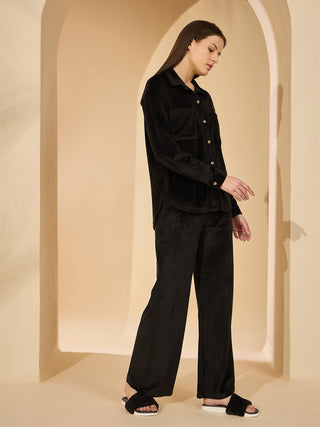 A woman wears a black velvet pajama set, featuring a long-sleeved button-up top with two chest pockets and matching wide-leg pants, accessorized with fluffy black slides.