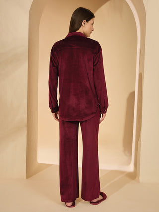 A woman in a burgundy velvet pajama set and slippers stands facing away from the camera, leaning against a beige wall.