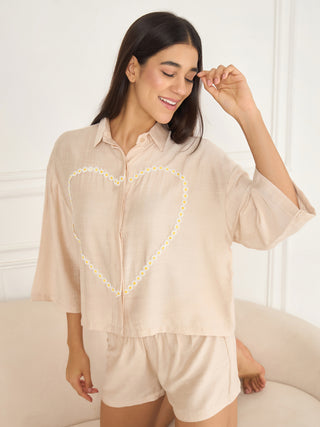 A woman poses playfully in a beige linen sleep set featuring a heart-shaped daisy print, a slight variation on the previous image with a different pose.