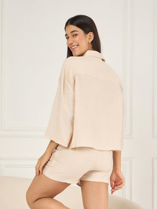 A woman wears a light beige linen pajama set consisting of a collared, cropped, oversized top with short sleeves and matching high-waisted shorts.