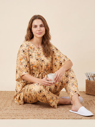 A young woman wearing a peach-colored floral print pajama set with a tie waist, sitting on a woven rug and holding a mug.