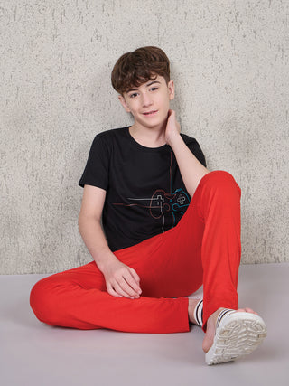 Boy seated on the floor against a textured wall, wearing a black short-sleeve T-shirt with multicolor gaming controller line print and red full-length pyjama pants.