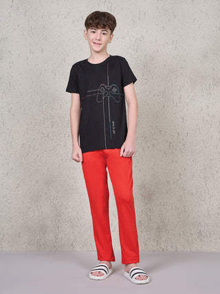 A young boy in a black t-shirt with a gamepad print and red pajama pants stands facing forward, wearing slides.