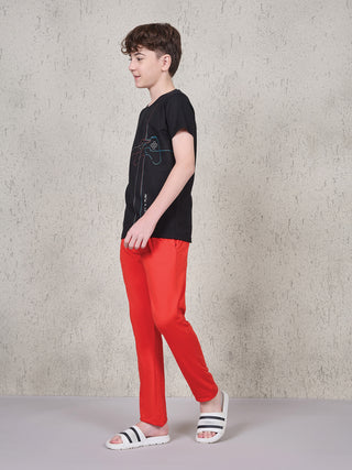 Boy standing in a side pose wearing a black short-sleeve T-shirt with multicolor gaming controller line print and red full-length pyjama pants.