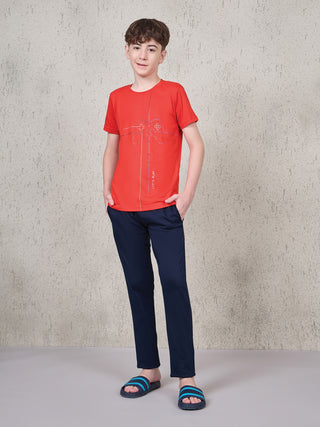 BOYS in a full-body standing pose wear a red t-shirt with a controller print and navy blue pants, with the addition of striped sandals.