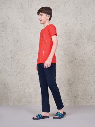 Boy standing in a side pose wearing a ferry red short-sleeve T-shirt with gaming controller line print and navy full-length pyjama pants.
