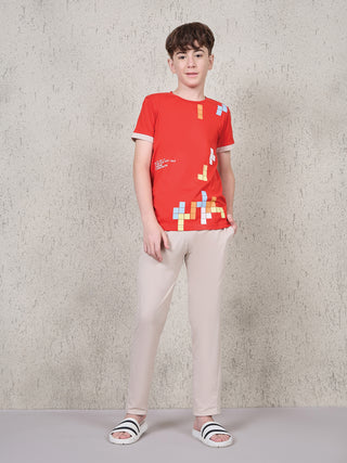 Boy standing front-facing with one hand in pocket, wearing a ferry red short-sleeve T-shirt with multicolor block print and beige full-length pyjama pants.