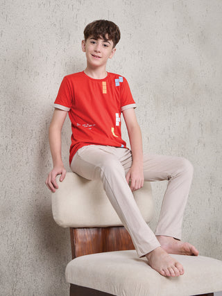 Boy seated on a beige chair wearing a ferry red short-sleeve T-shirt with multicolor block print and beige full-length pyjama pants.