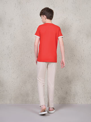 BOYS: A young boy is standing facing away from the camera, wearing a red short-sleeved t-shirt with light tan trim on the sleeves and light tan pants.