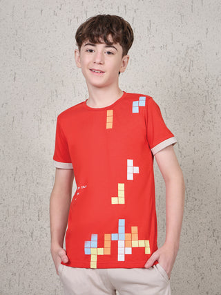 BOYS: A young boy stands facing the camera, wearing a red short-sleeved t-shirt with light tan trim on the sleeves and light tan pants, the t-shirt features a pixelated game-like print.