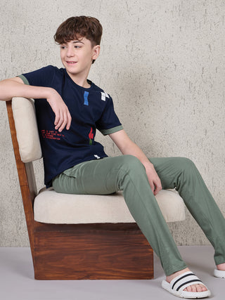 Boy seated on a beige chair in a relaxed pose, wearing a navy short-sleeve T-shirt with multicolor block print and olive green full-length pyjama pants.