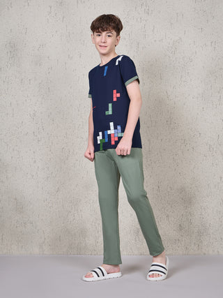 BOYS standing, wearing a navy blue t-shirt with a colorful pixelated print and olive green pants.