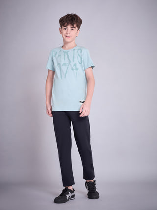 BOYS in a studio shot, wearing a light blue t-shirt with a graphic print and black pants.