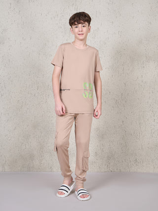 BOYS, standing, wearing a light brown t-shirt with neon green graphic print and light brown cargo pants, with white slides.