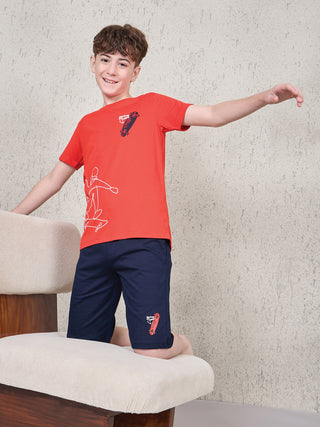 Boy kneeling on a beige chair in a playful pose, wearing a ferry red short-sleeve T-shirt and navy shorts sleepwear set with skateboard graphic print.