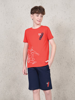 Boy standing front-facing with one hand in pocket, wearing a ferry red short-sleeve T-shirt and navy shorts sleepwear set with white skateboard graphic print.