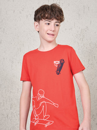 Boy standing in a slight side pose wearing a ferry red short-sleeve T-shirt with white skateboard graphic print and chest skateboard detail.