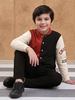BOYS wearing a color-blocked varsity jacket with cream, red, and black panels, and black pants, sitting on striped flooring.
