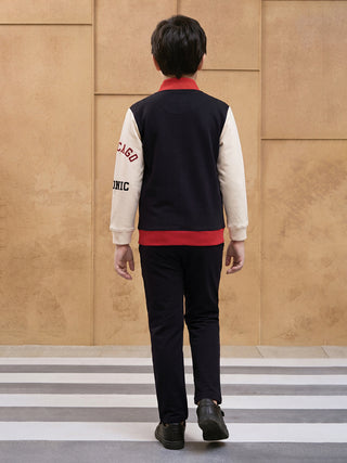BOYS wearing a navy and cream varsity jacket with red accents and black pants, walking away from the camera.