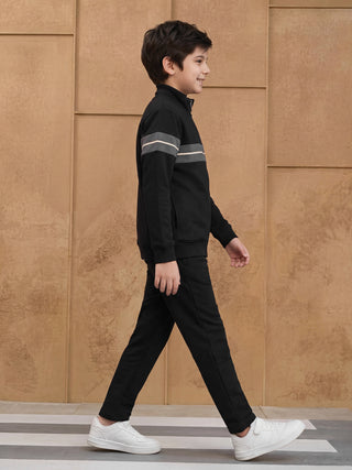 BOYS, A young boy walks from left to right wearing a black tracksuit with grey and white stripes on the sleeves and white sneakers.
