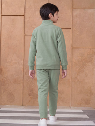 BOYS, wearing a light green fleece tracksuit consisting of a zip-up top and joggers, stands with his back to the camera, walking forward on a striped rug.