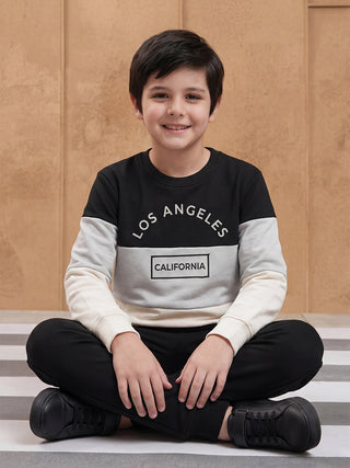 BOYS sitting on the floor with crossed legs, wearing a black, grey, and cream color-block sweatshirt with "LOS ANGELES CALIFORNIA" print and black sweatpants.