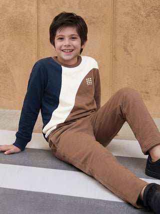 BOYS in a studio pose, wearing a brown and cream color-blocked sweatshirt with a subtle logo and matching brown sweatpants, carrying a tan duffel bag.