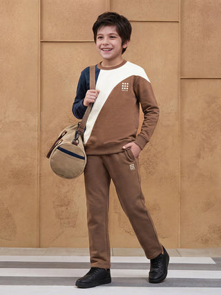 BOYS in a studio pose, wearing a brown and cream color-blocked sweatshirt with a subtle logo and matching brown sweatpants, carrying a tan duffel bag.