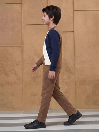 A young boy walks to the right wearing a color-blocked sweatshirt with navy blue, white, and brown panels, and brown trousers.